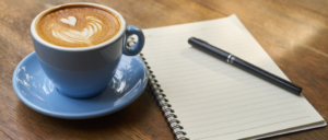 A blue mug with a frothy latte inside, next to a blank lined notebook and black pen
