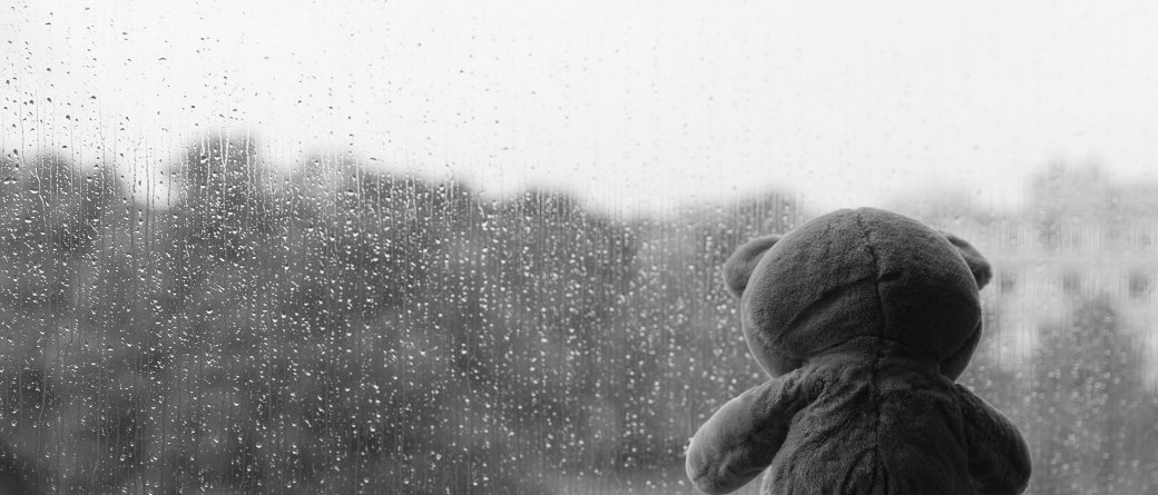 A greyscale image of a teddy bear facing a window covered in rain