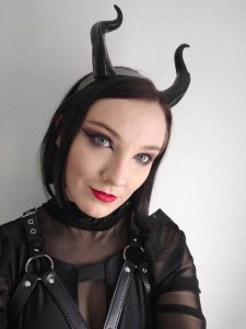 Photo of short story writer Isabella Hunter, who is wearing black clothes, red lipstick, and curved black devil horns. She has black hair.